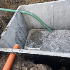 Szambo betonowe 10m3 cena z montażem. Betonowy zbiornik w wykopie z zieloną i pomarańczową rurą.