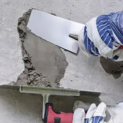 Naprawa betonu na zewnątrz: ręce w rękawicach nakładają zaprawę kielnią na uszkodzoną powierzchnię.