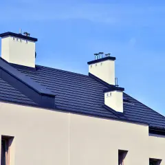 Nowoczesny dach bezokapowy z ciemnogranatową dachówką i białymi kominami, pod błękitnym niebem.