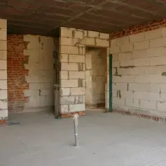 Wnętrze w budowie, ściany z bloczków betonowych i cegły, podłoga wylewana. Widać otwory drzwiowe i instalację hydrauliczną.