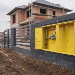 Żółta skrzynka z instalacją gazową w domu w budowie, obok ogrodzenia i wykopu z taśmą ostrzegawczą.