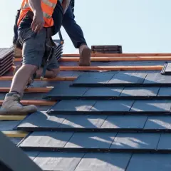 Pracownik w kamizelce odblaskowej układa dachówki. Trwa wymiana dachu, widać nowe materiały i drewniane łaty.