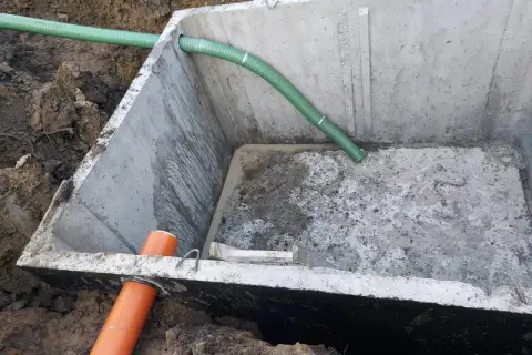 Szambo betonowe 10m3 cena z montażem. Betonowy zbiornik w wykopie z zieloną i pomarańczową rurą.