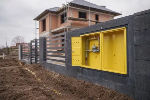 Żółta skrzynka z instalacją gazową w domu w budowie, obok ogrodzenia i wykopu z taśmą ostrzegawczą.