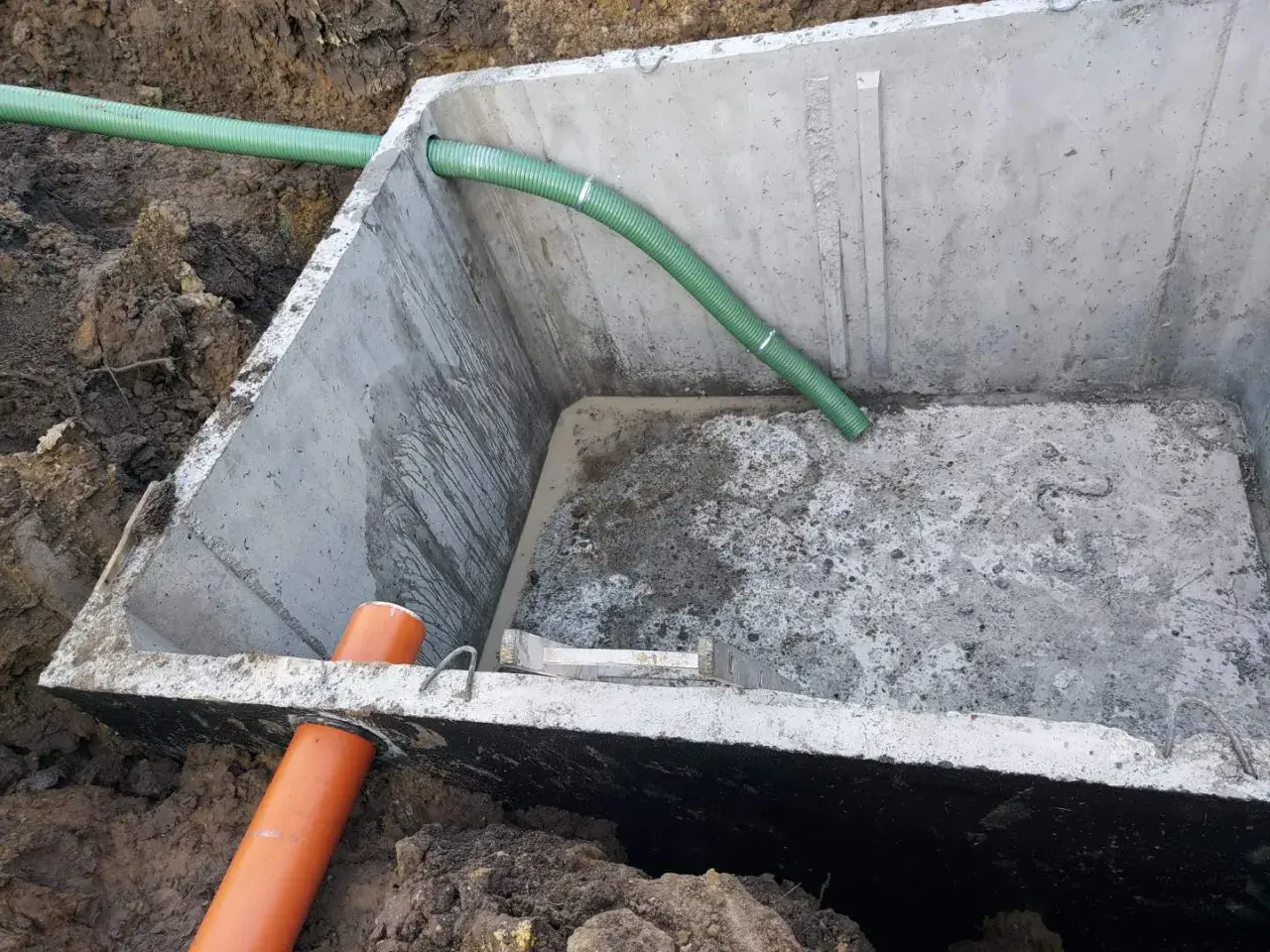 Szambo betonowe 10m3 cena z montażem. Betonowy zbiornik w wykopie z zieloną i pomarańczową rurą.