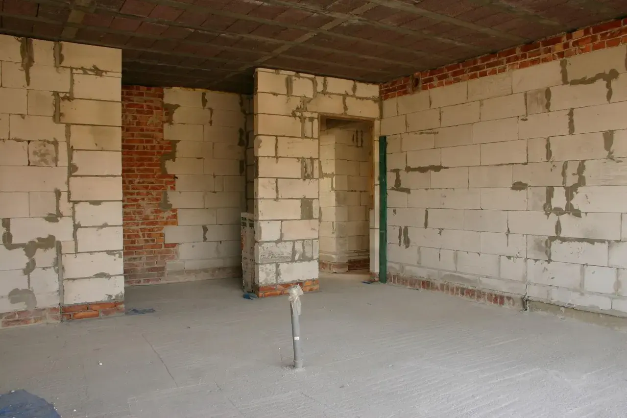 Wnętrze w budowie, ściany z bloczków betonowych i cegły, podłoga wylewana. Widać otwory drzwiowe i instalację hydrauliczną.