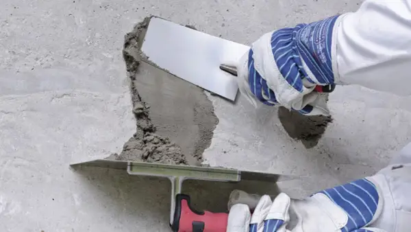 Naprawa betonu na zewnątrz: ręce w rękawicach nakładają zaprawę kielnią na uszkodzoną powierzchnię.