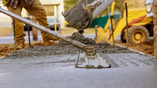 Pracownik wyrównuje świeży beton c12/15 za pomocą łaty. W tle widać koparkę i plac budowy.